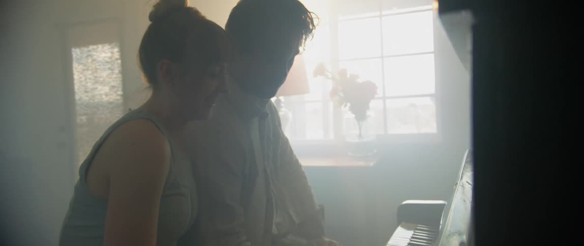 A man and a woman sit in front of a piano in a brightly lit room