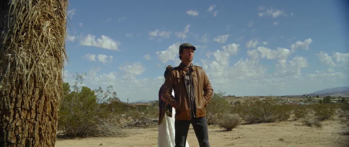 A man standing in a dusty and weed filled field while a woman wearing a long dress walks to him