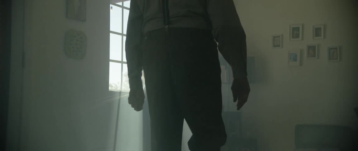 A man walks towards a lamp which stands under the window through which light streams in