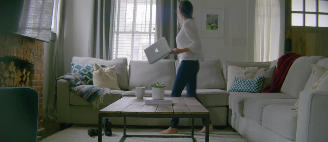 A woman is walking towards the couch with her laptop