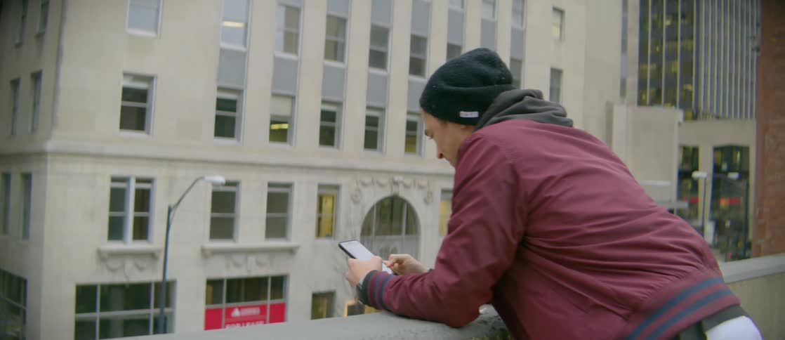 A man on a balcony overlooking a city building typing on his phone