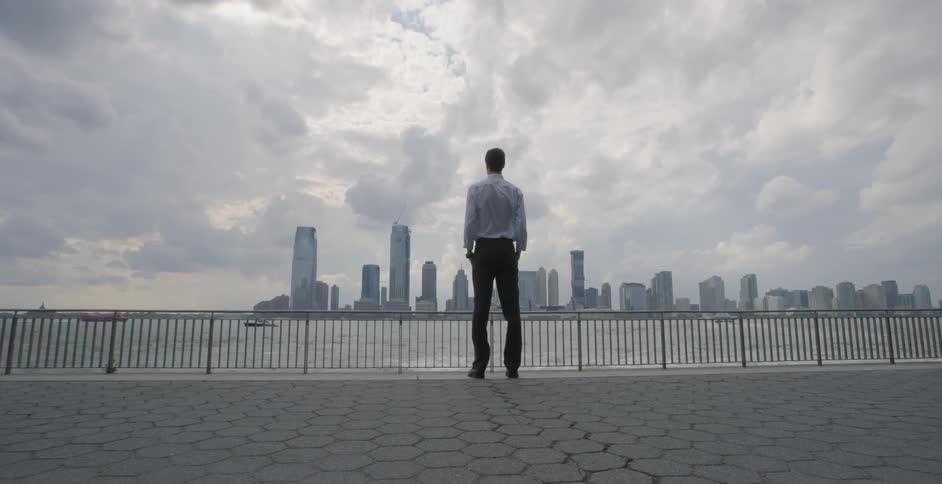 A man in a suit standing by river walkway staring at a city skyline