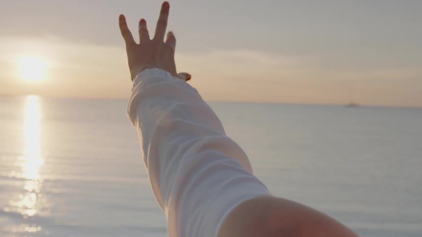 A woman on a beach reaches towards the setting sun