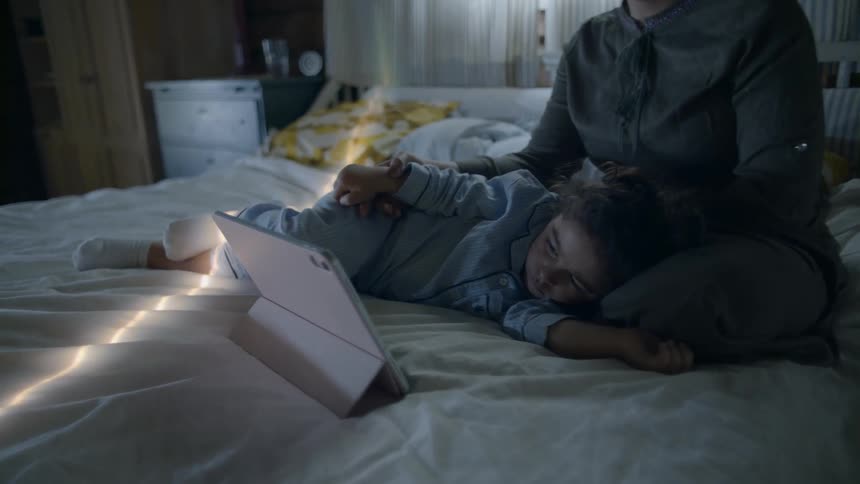 A woman and child on a bed watching a tablet
