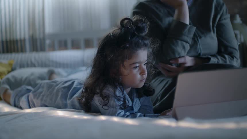 A little girl lies on a bed beside her sitting mom while watching a video