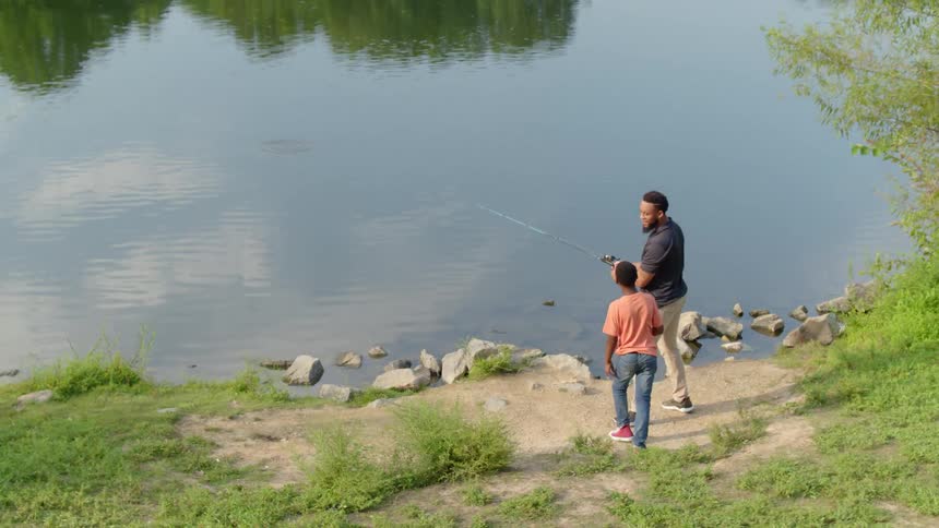A boy stands next to his dad as he fishes in a lake on a sunny day