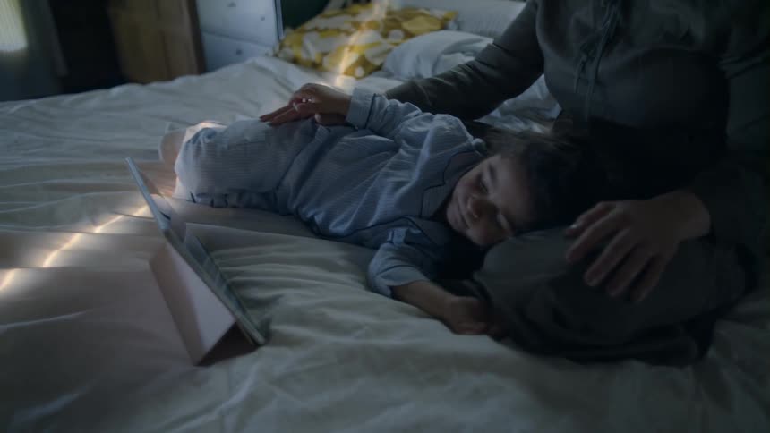 A woman sit on her bed with her child sleeping in her lap as she watches a video