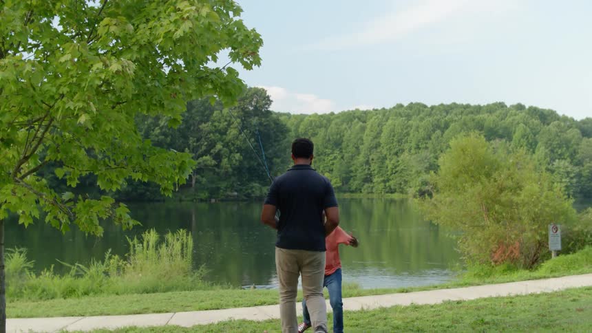 A dad and his son carry fishing poles down to a pond