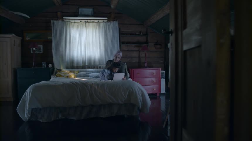 A mother and young daughter use a tablet in bed