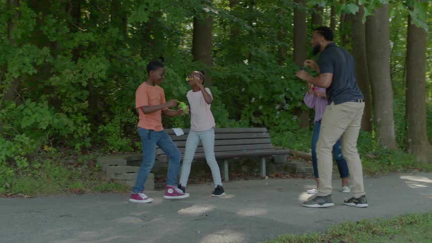 A black family blows bubbles in a heavily wooded park