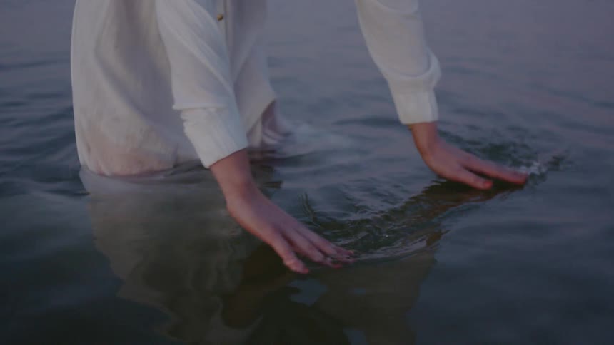 Woman wearing a white dress swishing her hands through water she is standing in