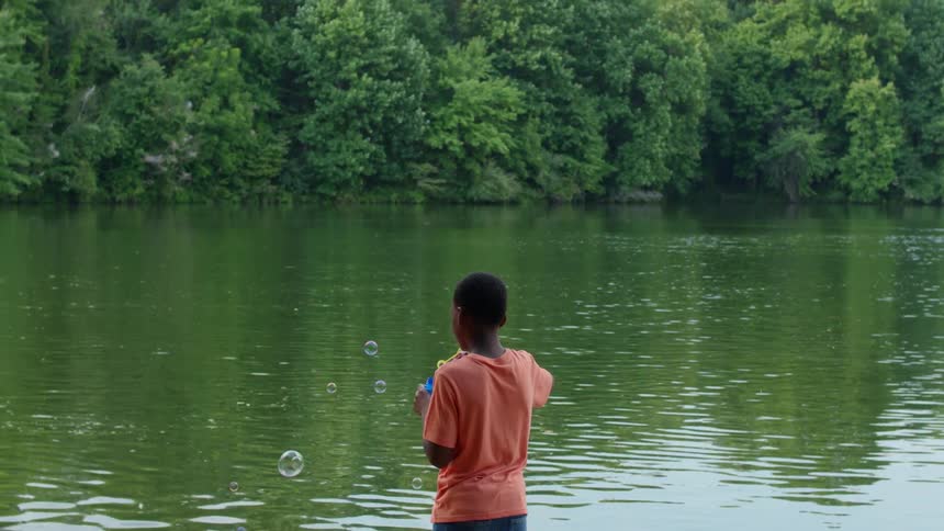 A boy is outside near a lake blowing bubbles