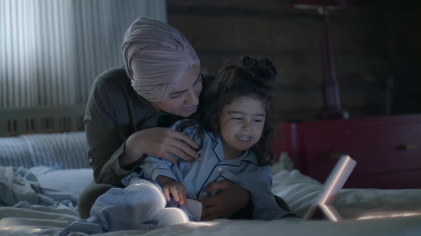 Mother and child in pajamas sit on bed and look at tablet