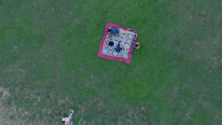 A child runs with a kite away from the blanket in the grass where his family lies and sits