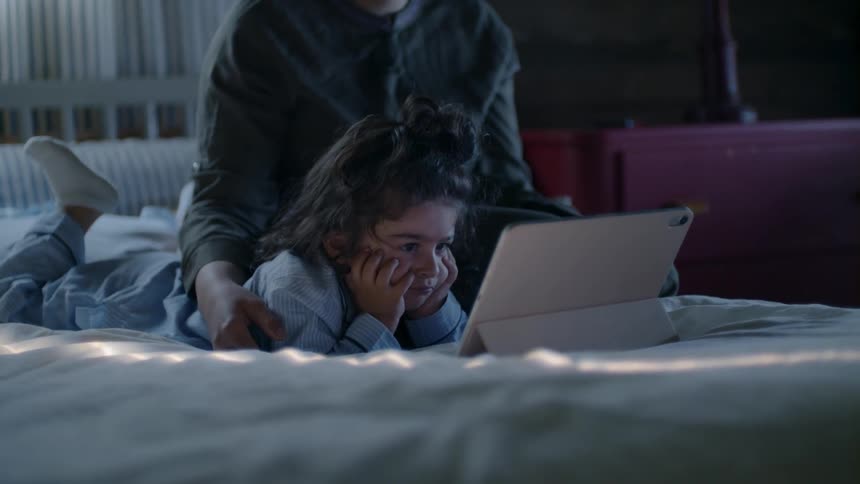 A child laying in bed watching a tablet with a woman