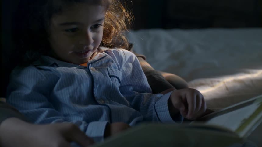 A little girl sitting on a bed and reading a book with another person