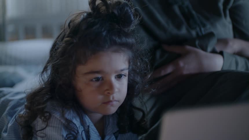 A girl in pajamas watches a tablet while in bed
