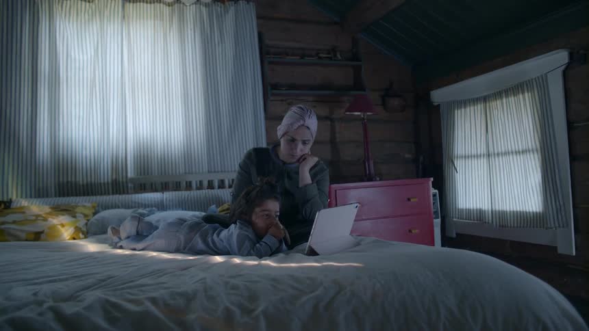 A mother and daughter look at a tablet in bed