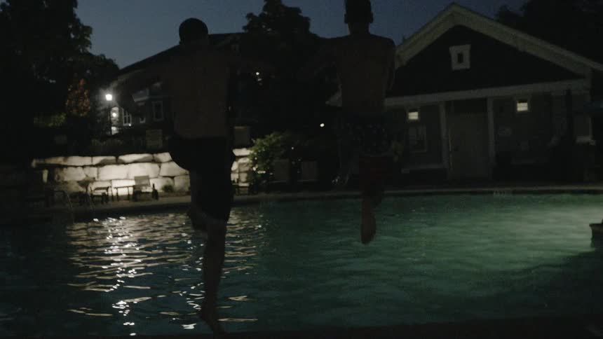 Two men jump in the pool together at night