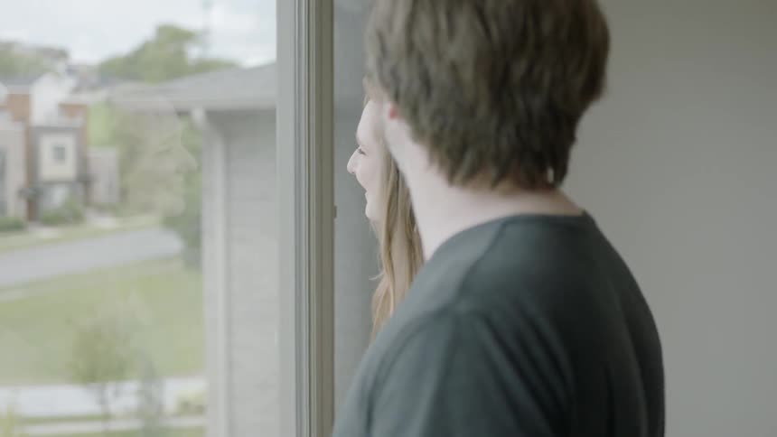 A man and woman walk to a window and smile as they look out over the neighborhood