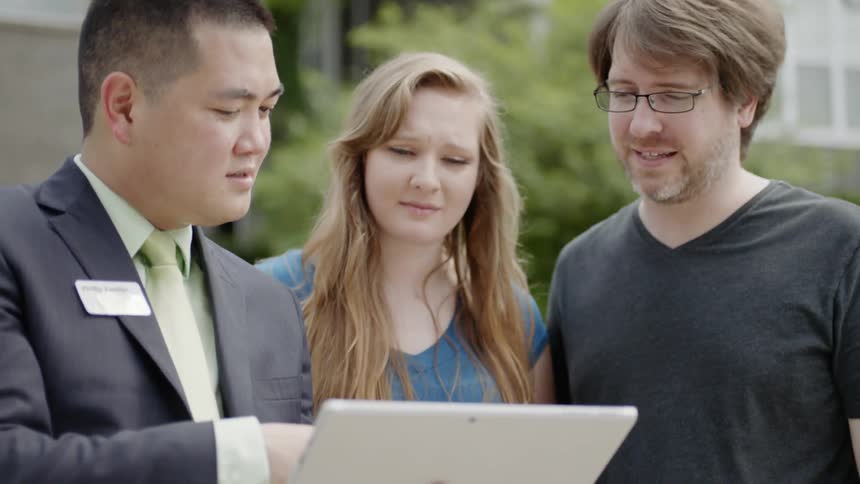 Man wearing suit points out houses and show information to man and woman on a sunny day