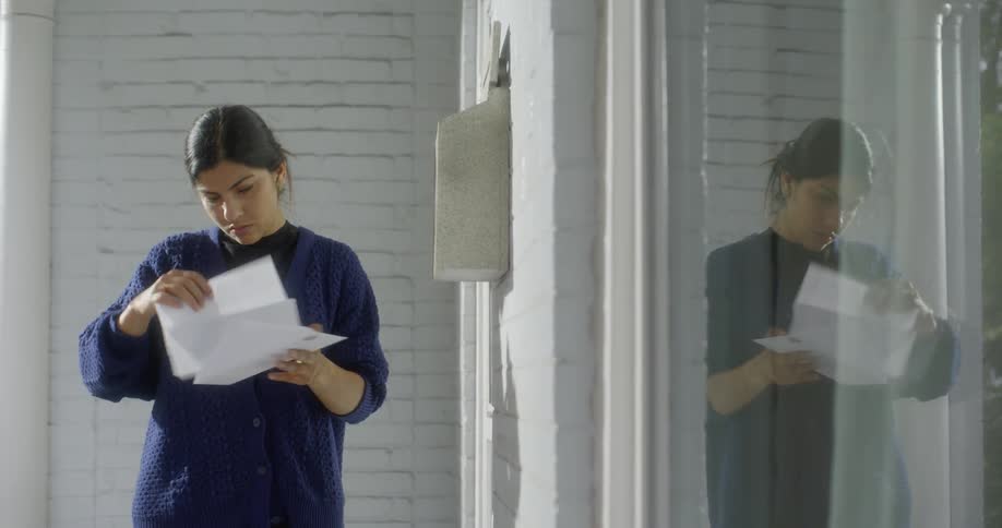 A woman checks her mailbox and drops things on the ground in her rush to open a letter with a picture