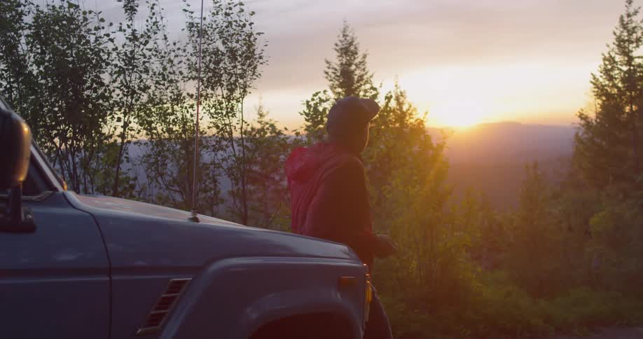 A man leaning on a jeep looking at a sunset in the forest
