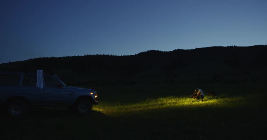 A man and his dog are in the glow of the headlights of a truck on grassland at sunset