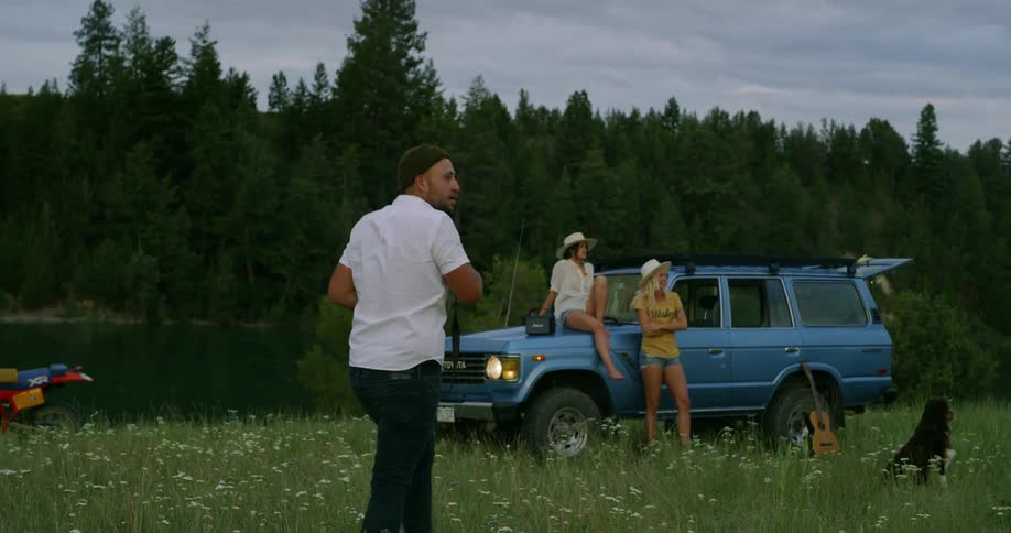 Man takes picture of two women on a blue car next to dog by lakeside in forests a cloudy day