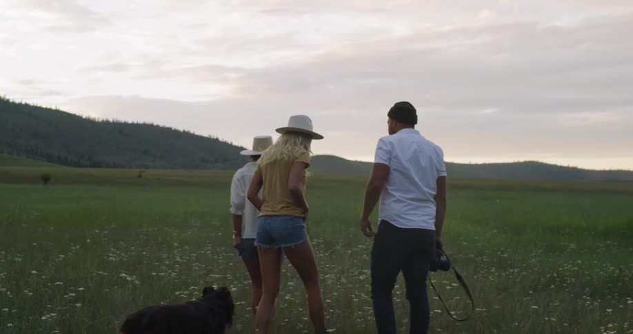 A man and two women walk with a dog through a meadow until the man takes a photo of the remaining three