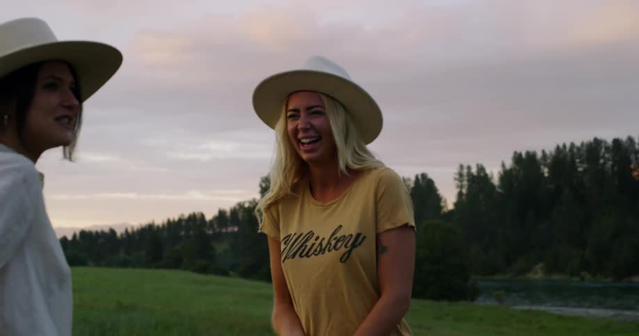 Two women wearing large hats standing in a field by a river while laughing