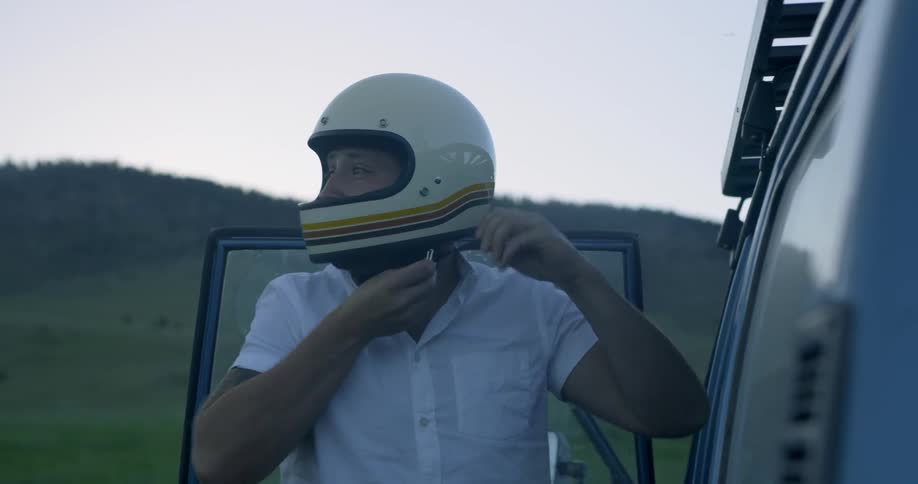 Man who wears white shirt put on motorcycle helmet on green field by dusk