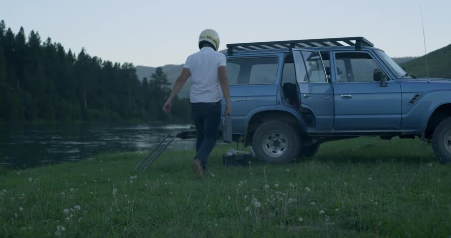 A man in a helmet walks up to a car parked next to a rural river