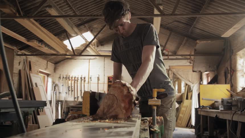 A young man blowing sawdust off a saw table before setting a portion of a log on it and sawing it