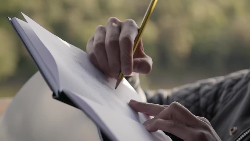 A person writes in a notebook using a pencil while outdoors