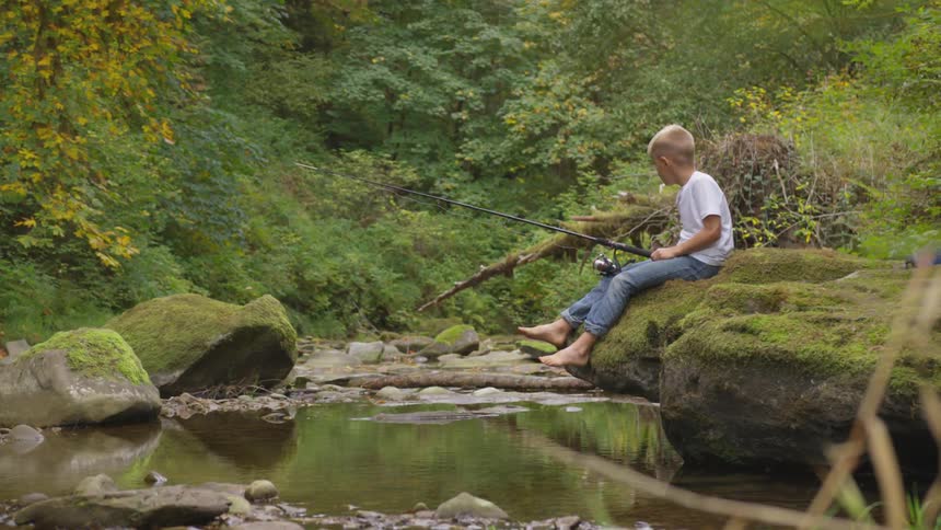 Boy sitting on a mossy rock fishes in a stream
