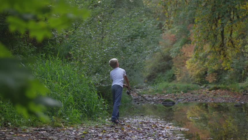A young boy casts a fishing pole into a small lake while he stands on the rocky shore