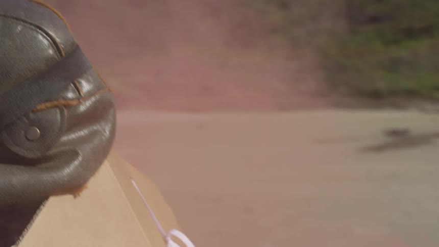 A child wearing  a green hat, goggles, and a cardboard cape is running on a dirt road with hills in the background