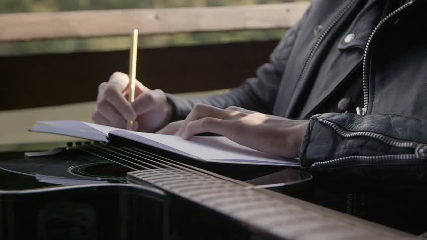 A person sits writing into a notebook with an acoustic guitar resting across their lap