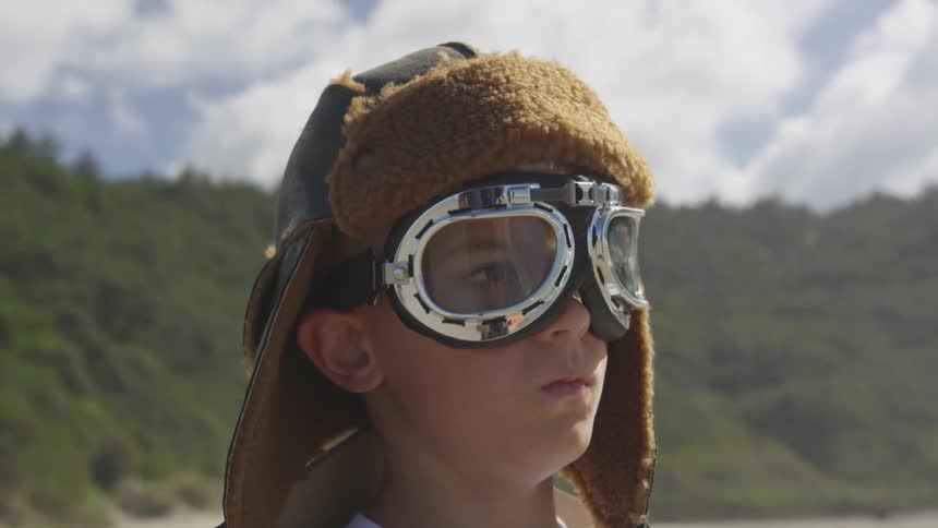 A boy wearing goggles and a hat with ear flaps stands outside
