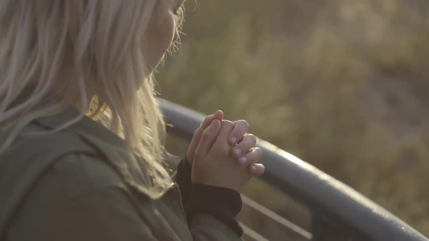 A woman stands outside by a railing praying with clasped hands