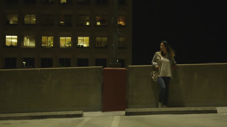 A woman leans against the wall in the parking lot at night