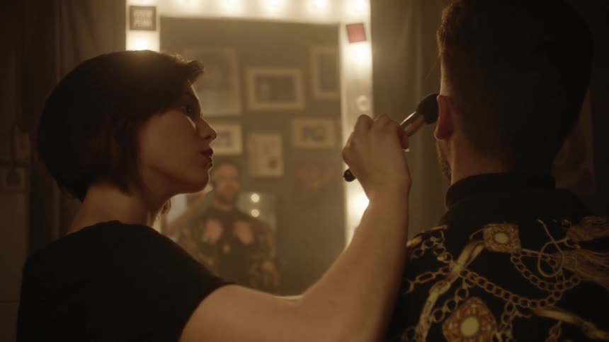 A makeup artist applies makeup to a man in a dressing room