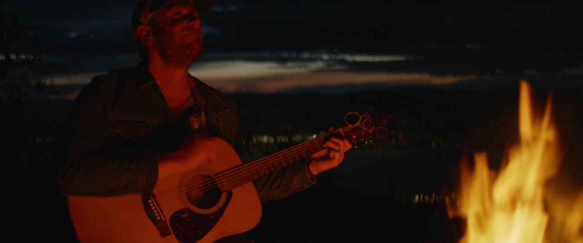 A man wearing a baseball cap strums a guitar around a campfire