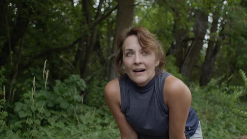 A woman is in a wooded area on a path leaning over and taking a breath