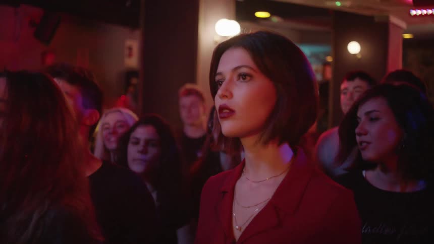 A group of young adults including a woman in a red top with a bob haircut sway to the music at a show