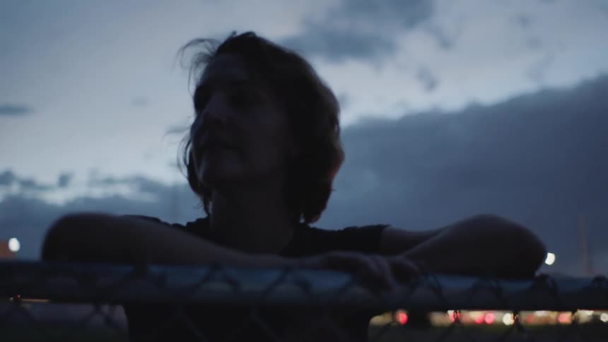Woman leans on a chain link fence at sunset