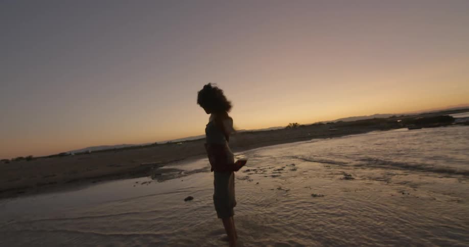 A woman wearing a sarong raises her arms and spins as she paddles in the sea at dusk