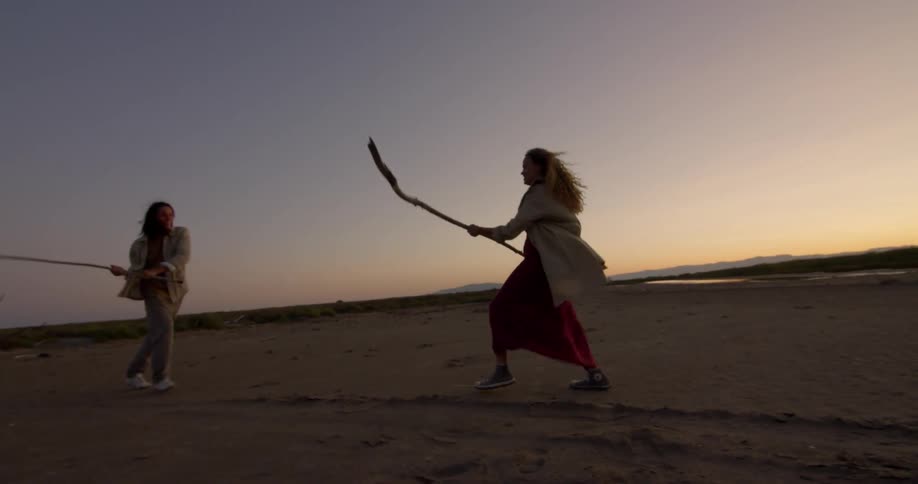 A man and woman playfully sword fight with long branches on a beach