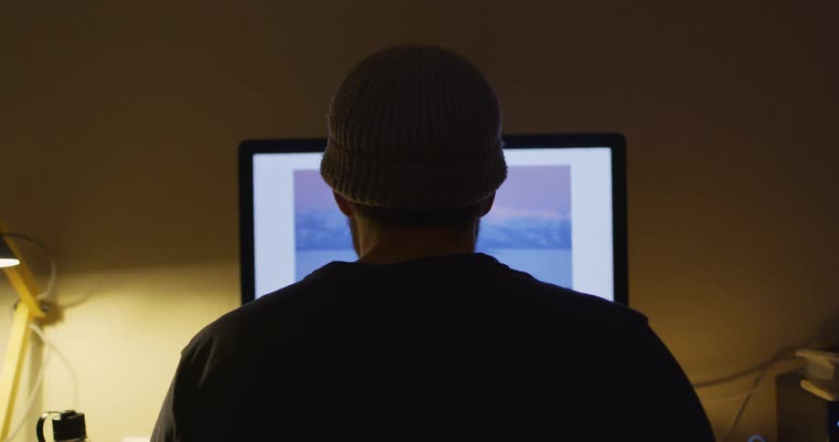 A man in a beanie hat looks at a photograph on a desktop monitor as a candle flickers